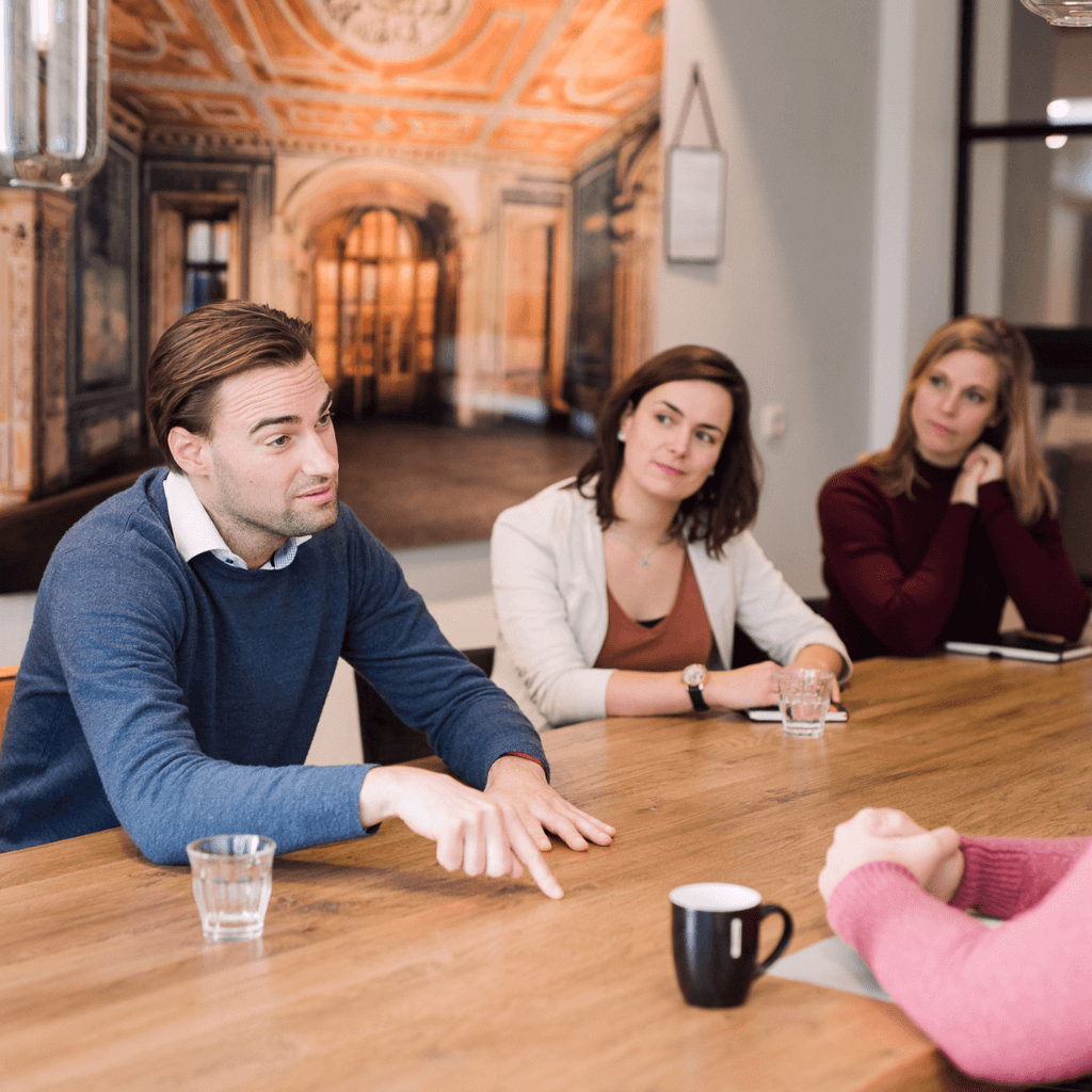 A man explaining something during an in-person meeting at a table who has fully attention of at least three other persons who are sitting around the table.