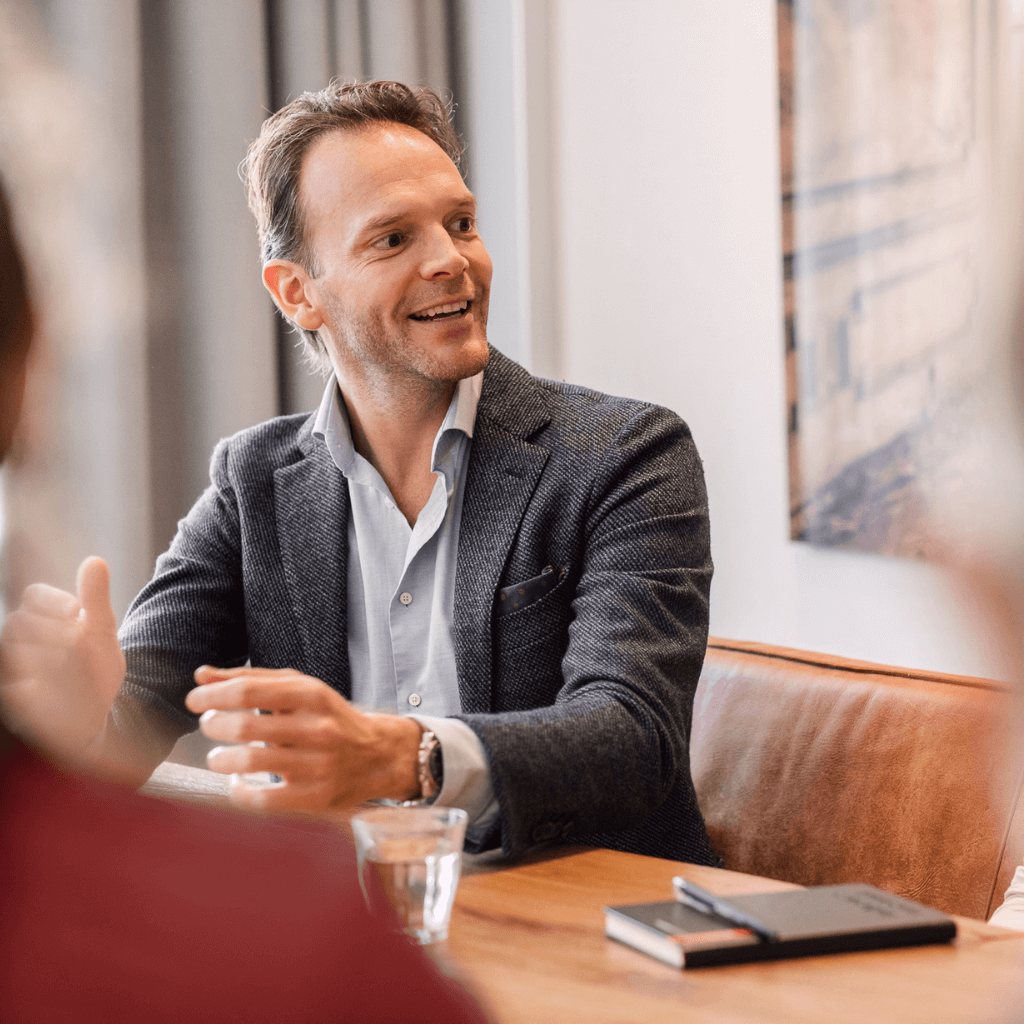 A man sitting on a chair to a table explaining something with a positive attitude by smiling and using his hands while talking to someone.
